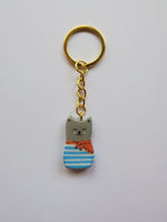 Porte-clé Lucky Rolie Cat Eddy — petit chat gris breton espiègle en marinière, modelé et peint à la main en argile, fini à la résine UV. Pièce unique fait main en Bretagne.